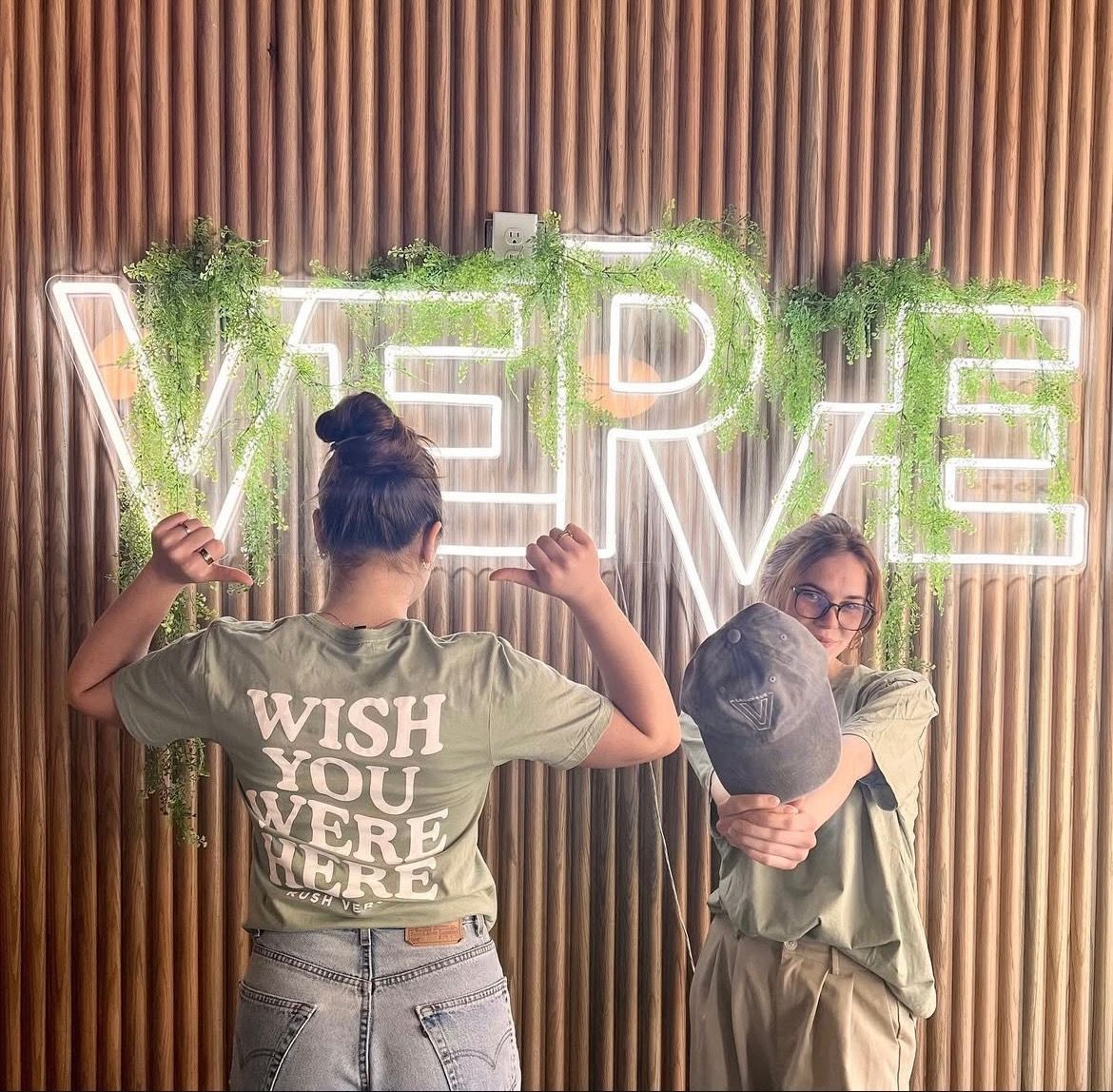 Two students posing in front of a glowing VERVE Columbus sign with hanging greenery—one pointing to the back of a “Wish You Were Here” shirt, and the other holding up a VERVE hat with a playful smile.