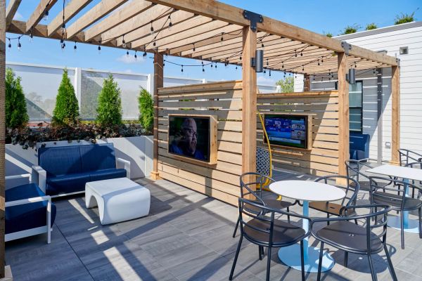 Lounge area at VERVE Columbus featuring a wooden pergola with string lights, mounted outdoor TVs, modern seating, and lush greenery—perfect for game day hangouts or relaxing with friends.