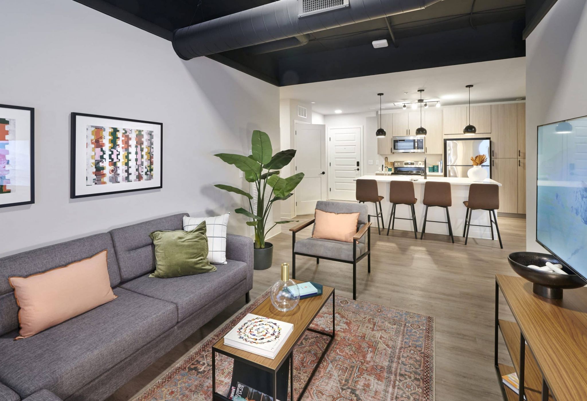 Modern apartment living room at VERVE Columbus featuring a gray sofa with accent pillows, a stylish coffee table, a large plant, and an open kitchen with bar seating under pendant lights.